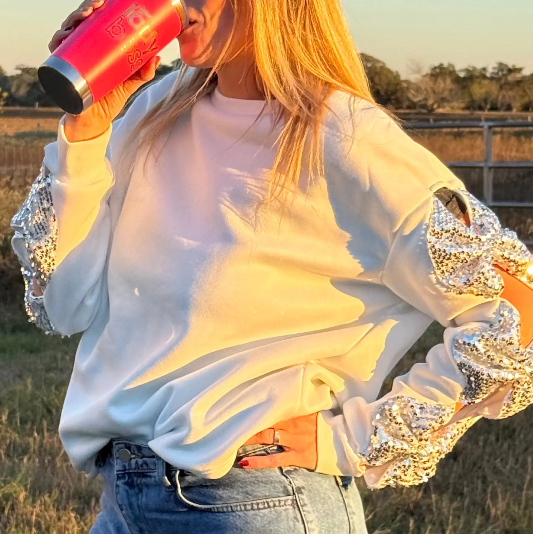 Person wearing a white sweatshirt with lace sleeves, drinking from a pink cup outdoors.