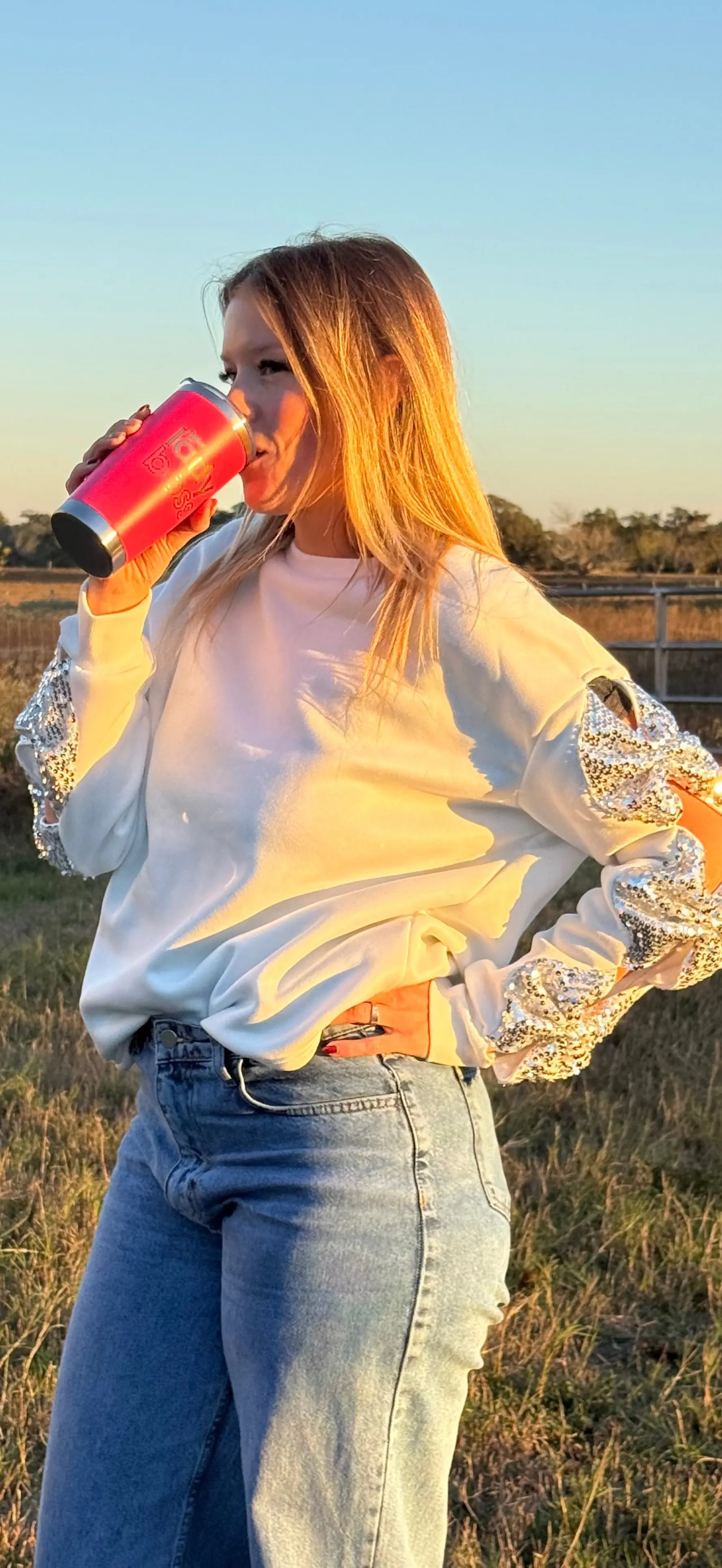 Person wearing a white sweatshirt with lace sleeves, drinking from a pink cup outdoors.